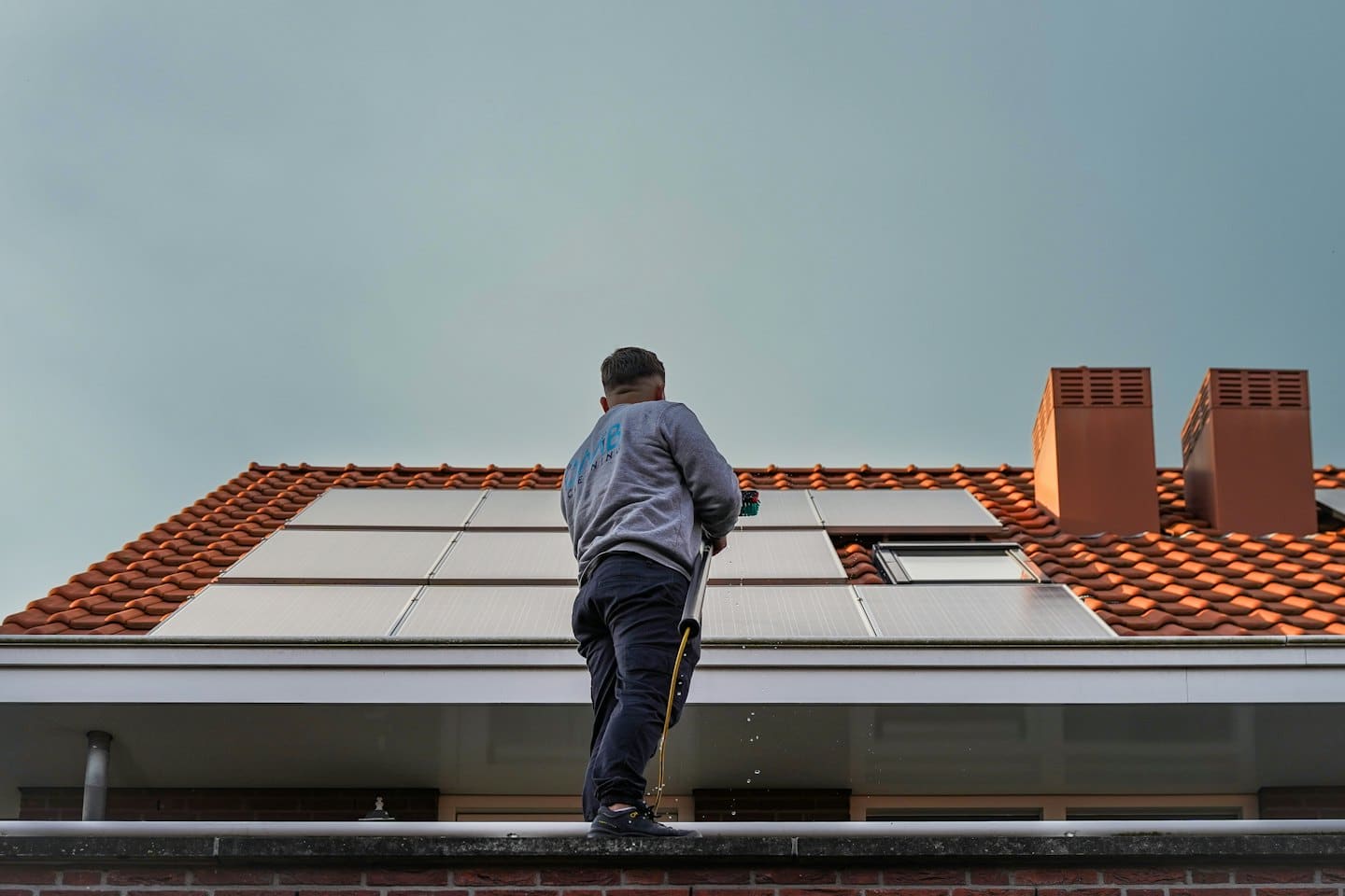 Werknemer maakt zonnepanelen schoon op een oranje pannendak met een lange borstel.